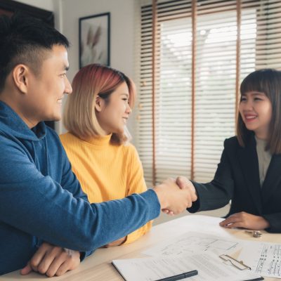 Happy young Asian couple and realtor agent. Cheerful young man signing some documents and handshaking with broker while sitting at desk. Signing good condition contract.